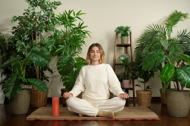 We. HEAR 1 coral red – tragbarer 40-Watt Bluetooth-Lautsprecher neben Frau beim Yoga vor Pflanzenhintergrund